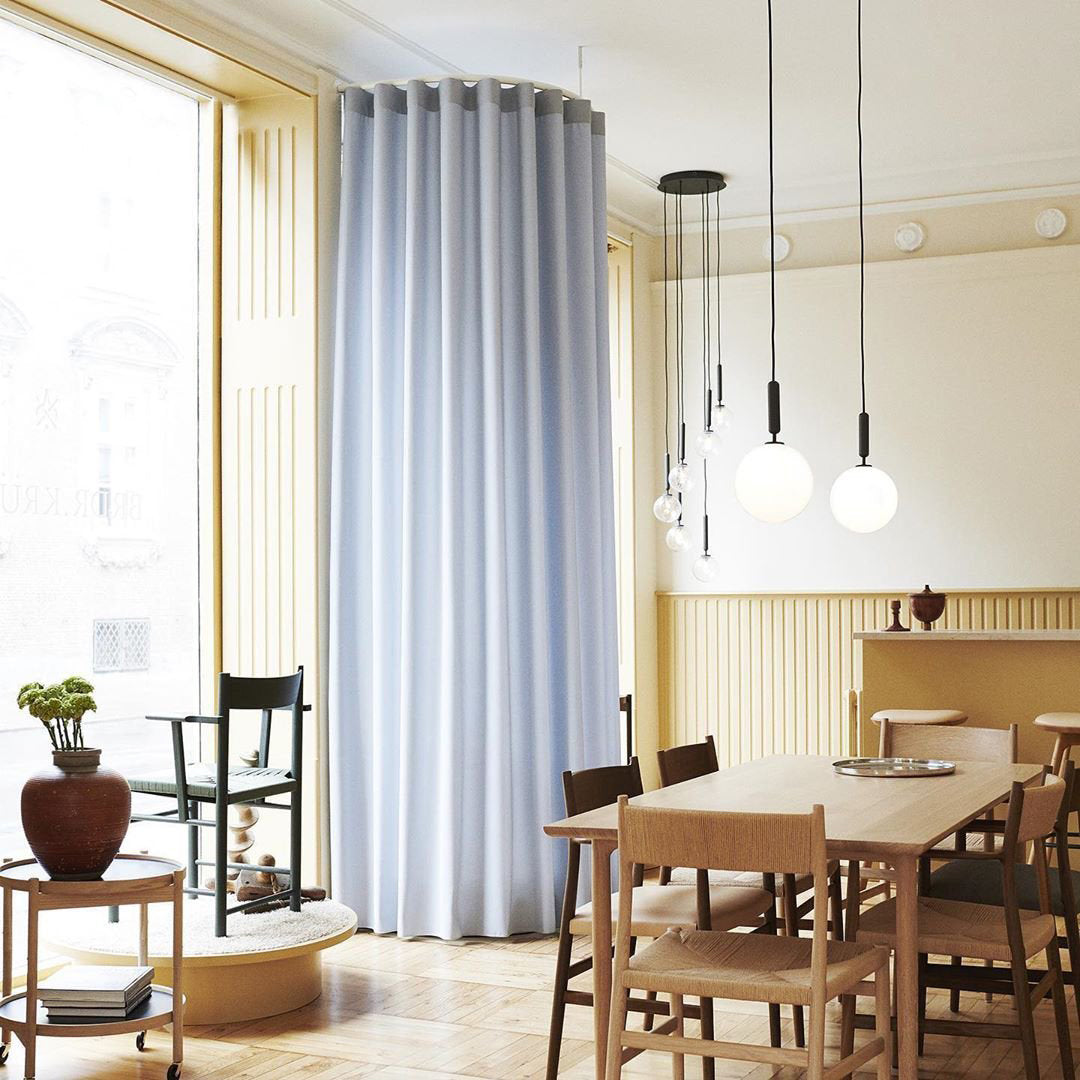 Several Clérachic pendants in brass and white opal glass hang over a dining table in a room with pale blue curtains.