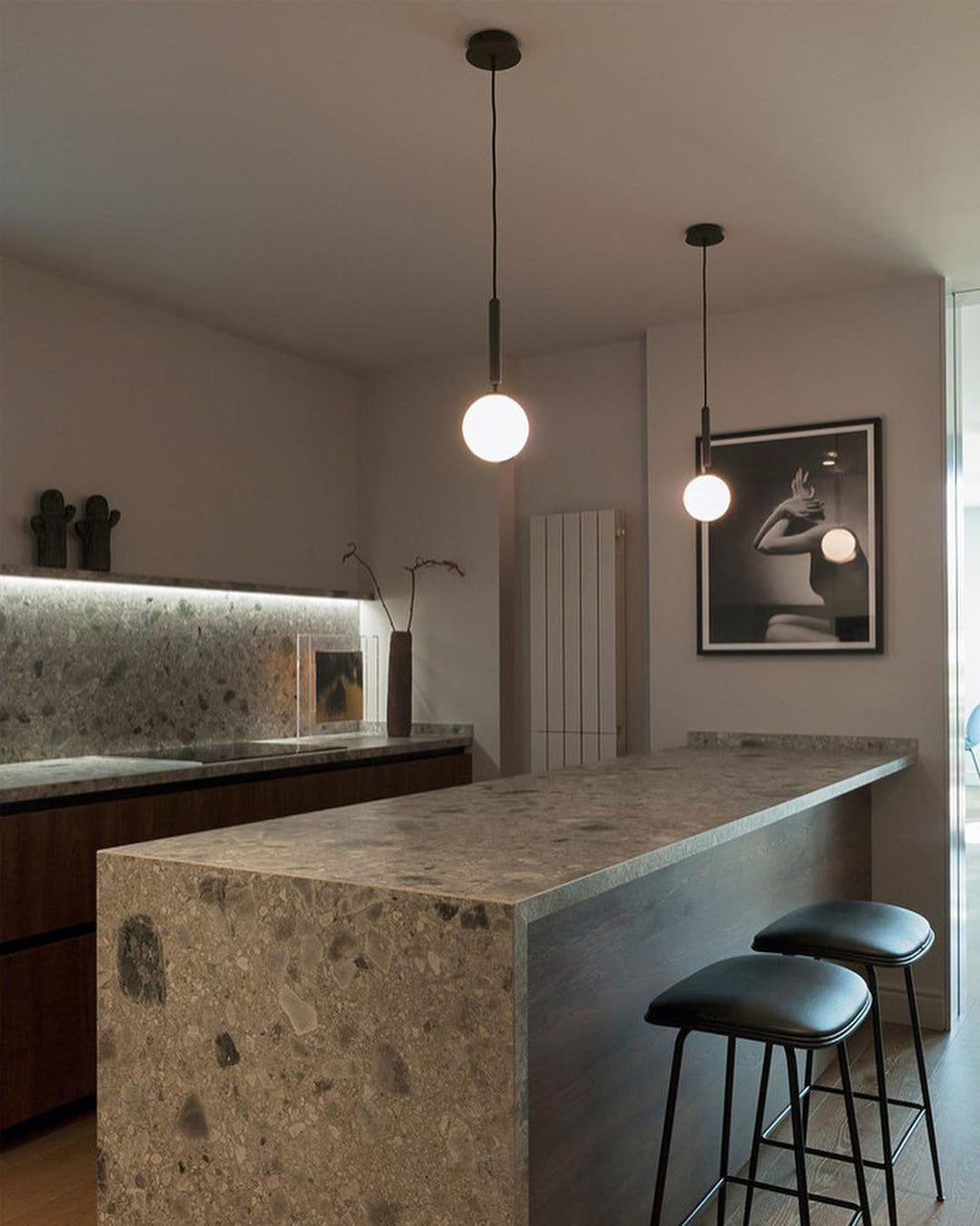 Two Clérachic pendants with white globes hang over a stone-topped kitchen island with black leather stools.