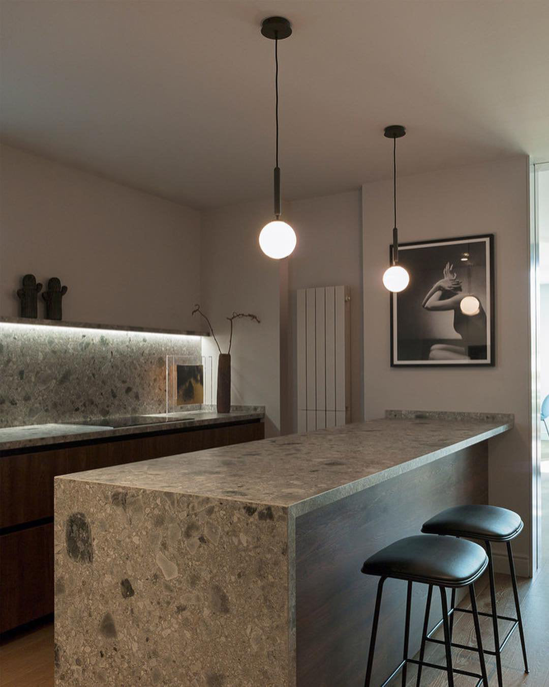 Two Clérachic pendants with white globes hang over a stone-topped kitchen island with black leather stools.