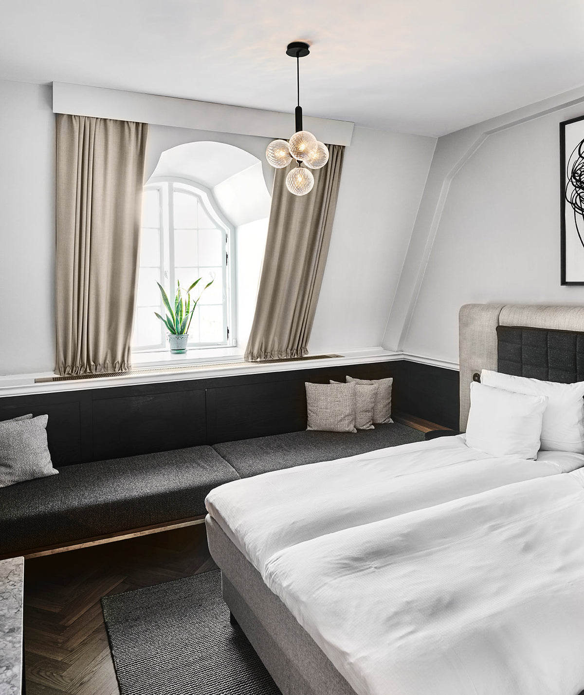 A Clérachic cluster pendant with four white globes hangs over a bed in a room with arched window and grey curtains.