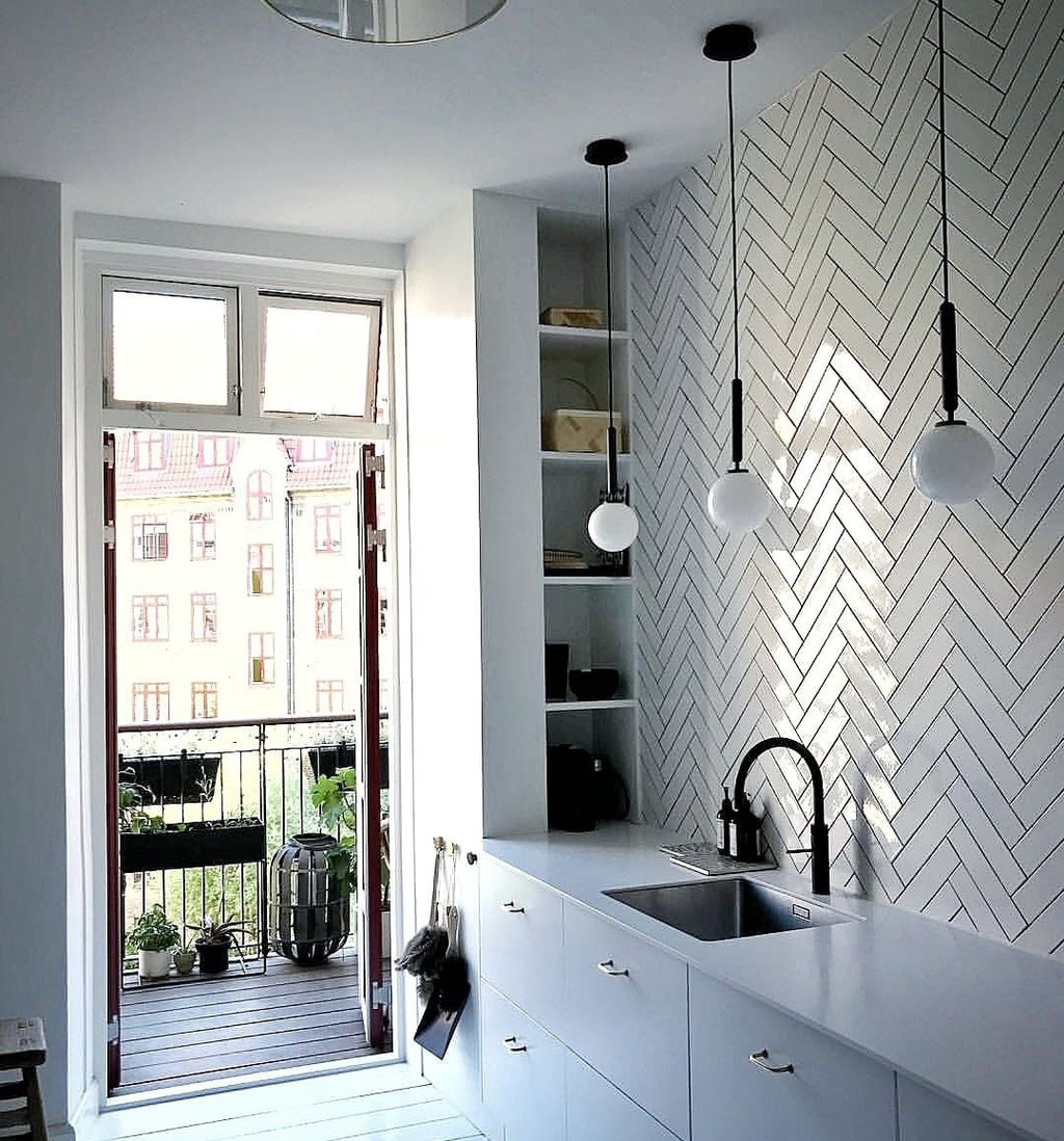Three single-globe Clérachic pendants on black rods hang over a kitchen counter with a herringbone tile wall.