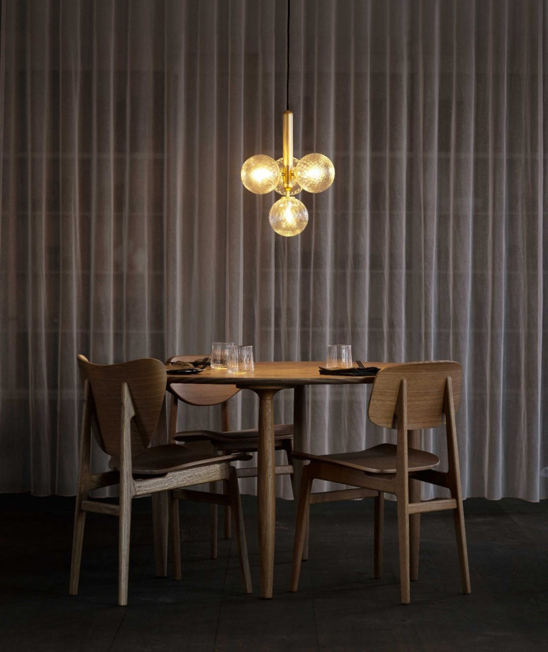 Brass cluster pendant from Clérachic glows amber above a round wooden table, with floor-length curtains behind.
