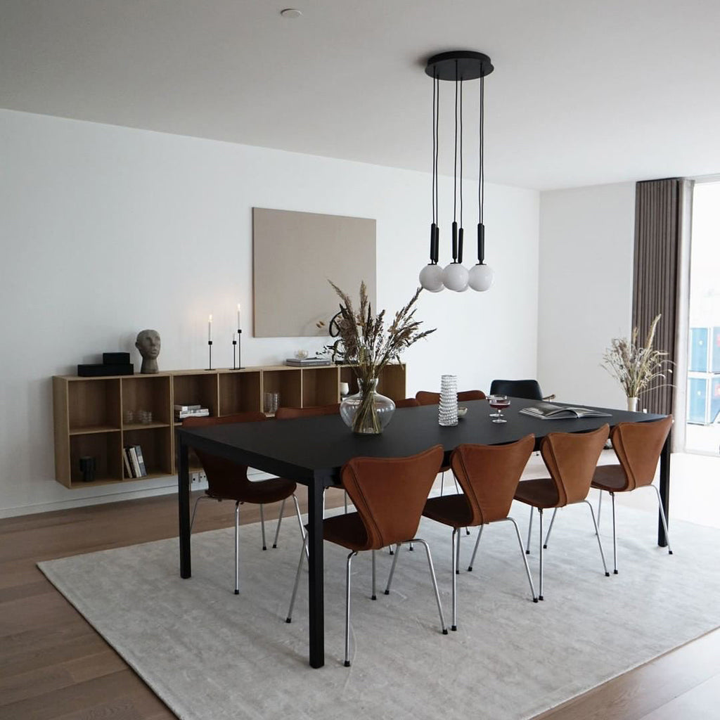 A round-plate Clérachic pendant with three white globes hangs above a black table, with oak shelves in the background.