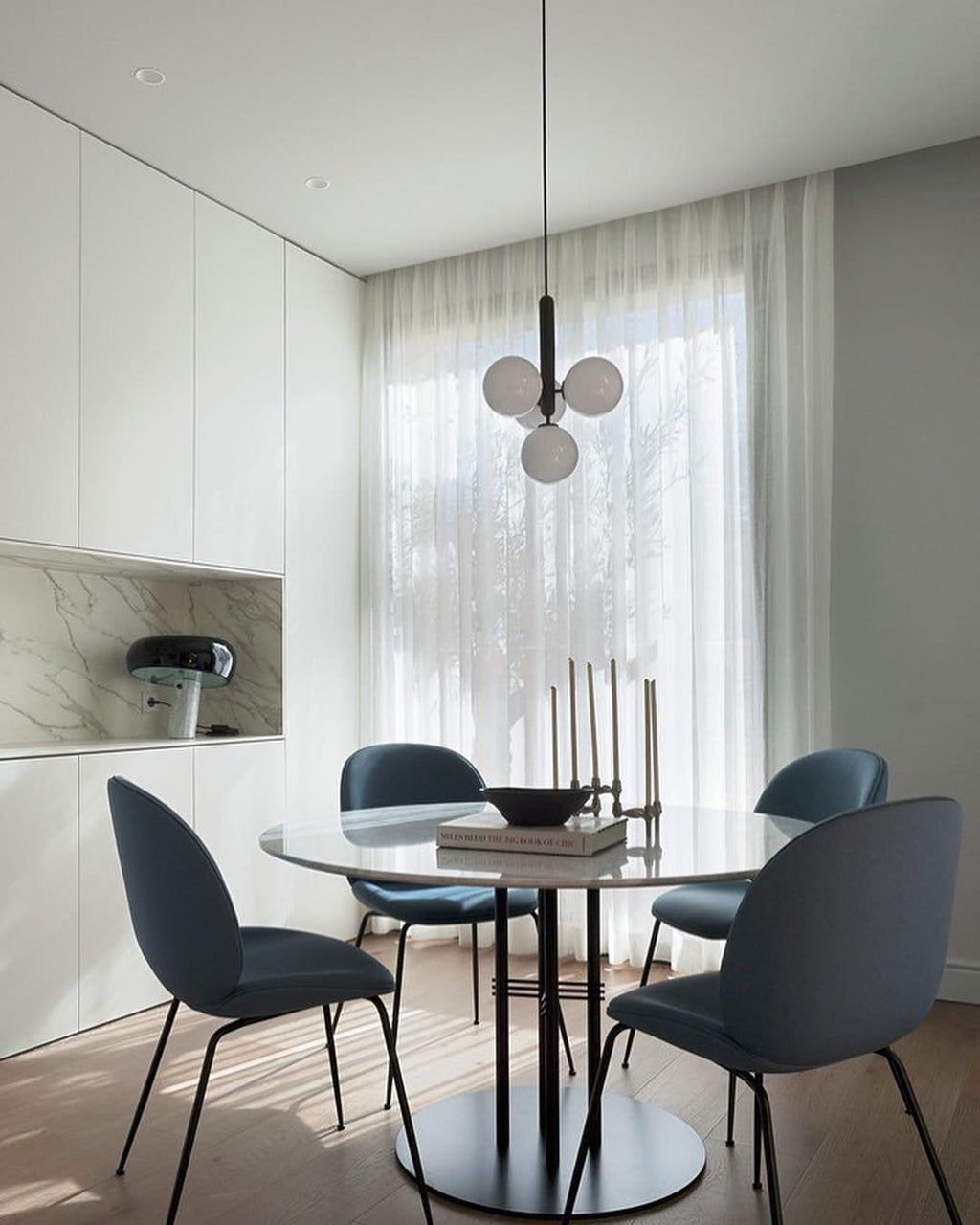 A Clérachic pendant with a black rod and white globes hangs between sheer curtains above a round table and blue chairs.