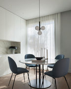 A Clérachic pendant with a black rod and white globes hangs between sheer curtains above a round table and blue chairs.