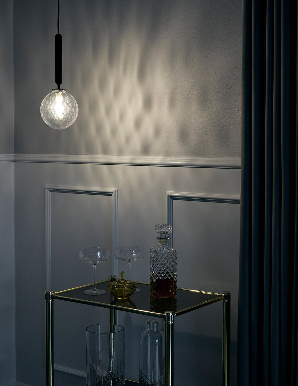 A single clear textured glass globe from Clérachic glows beside a dark wall, throwing a pattern of light.