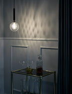 A single clear textured glass globe from Clérachic glows beside a dark wall, throwing a pattern of light.
