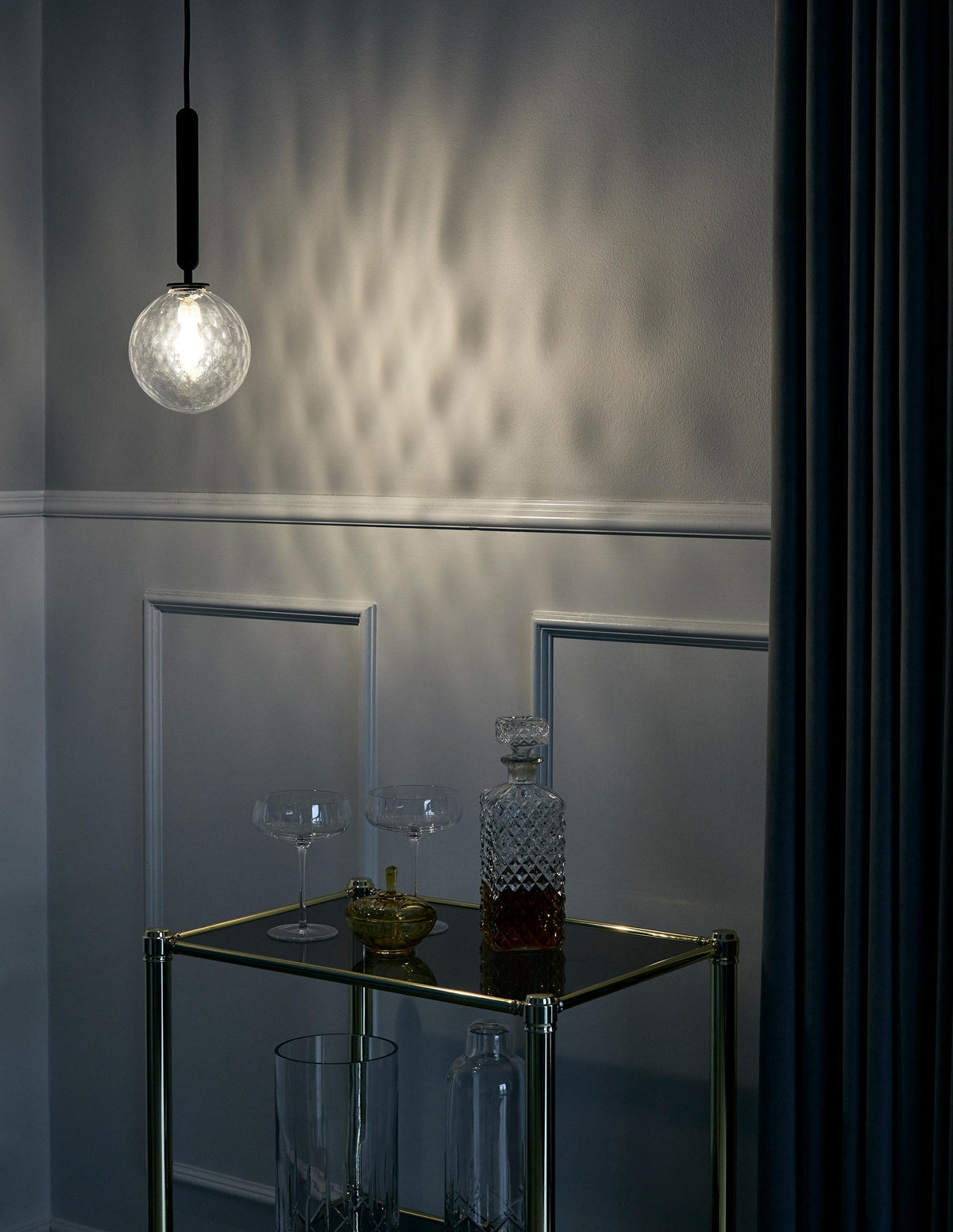 A single clear textured glass globe from Clérachic glows beside a dark wall, throwing a pattern of light.