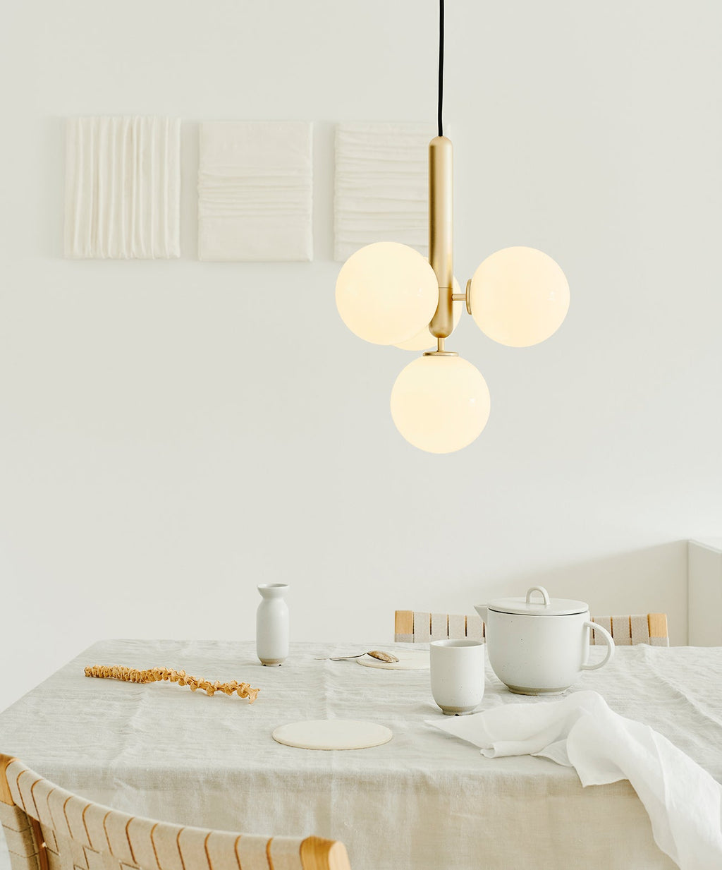 A brass-rod Clérachic pendant with three white globes hangs close to the wall above a table with pale ceramics.