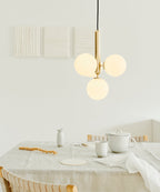 A brass-rod Clérachic pendant with three white globes hangs close to the wall above a table with pale ceramics.