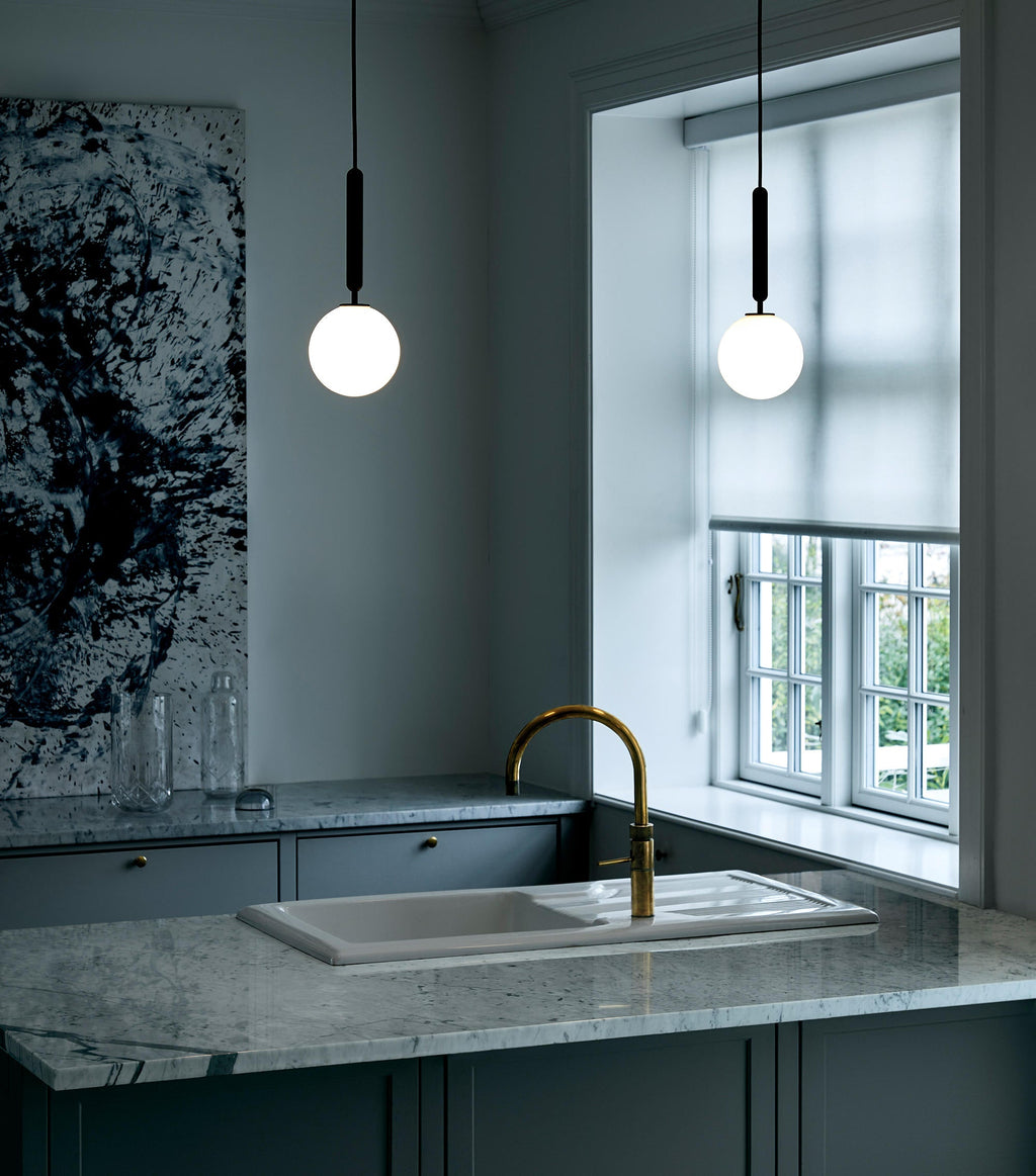 Two single-globe Clérachic pendants with black rods hang in a dark kitchen beside a window with a brass tap.