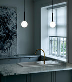 Two single-globe Clérachic pendants with black rods hang in a dark kitchen beside a window with a brass tap.