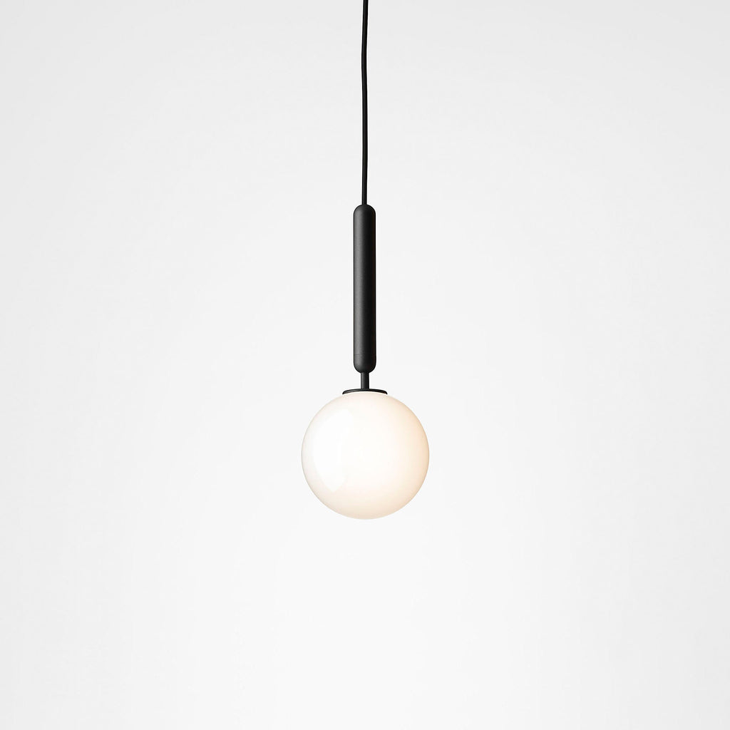 Single Clérachic pendant with a black rod and one white opal globe, lit softly against a pale grey backdrop.