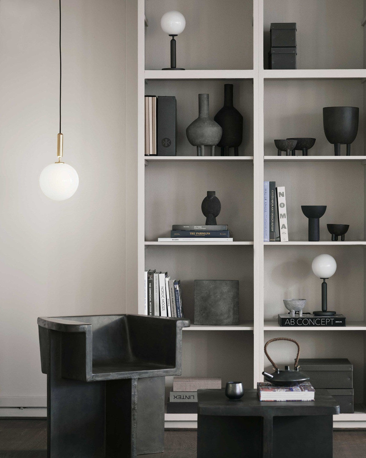 A single-globe Clérachic pendant on a brass rod hangs near a grey wall and a tall shelf with dark ceramics.