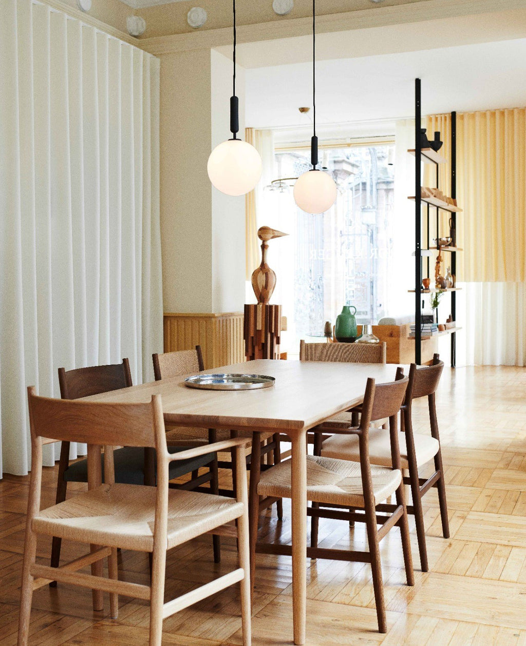 Two Clérachic pendants with white opal globes hang at different heights over a pale oak table and chairs.