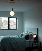 Clérachic white-globe pendant with a black rod hangs over a bed with teal linen and a small lamp beside it.