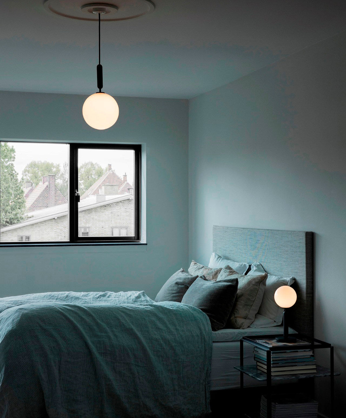 Clérachic white-globe pendant with a black rod hangs over a bed with teal linen and a small lamp beside it.