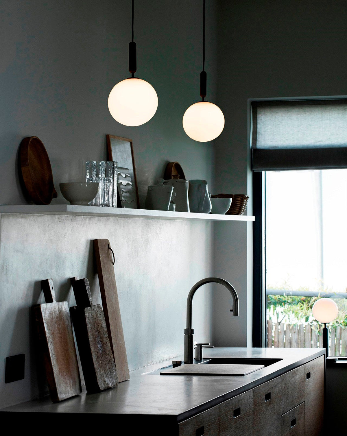 Two white-globe Clérachic pendants hang over a concrete kitchen counter with a sink and a window onto green.