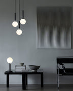 Three black-rod Clérachic pendants with opal globes hang at different heights above a low table and wall panel.