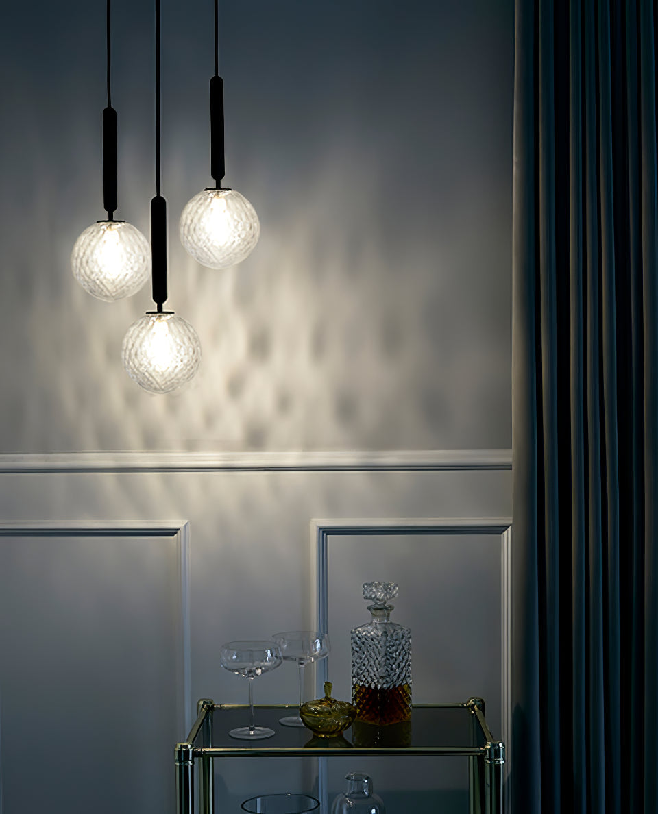 Three clear textured globes glow on this Clérachic pendant's black rods above a drinks trolley, casting a diamond pattern on a panelled wall.