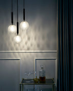 Three clear textured globes glow on this Clérachic pendant's black rods above a drinks trolley, casting a diamond pattern on a panelled wall.