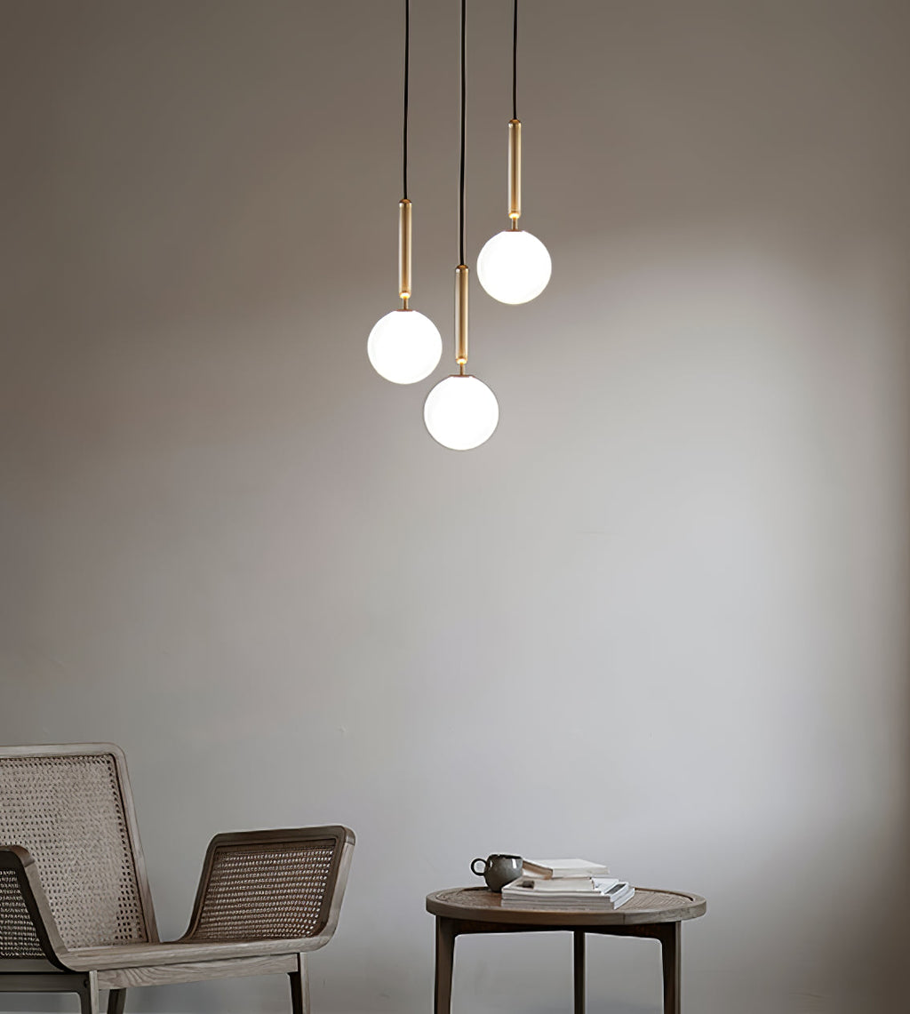 Three brass-rod Clérachic pendants with white globes hang in a loose group in front of a warm grey wall.
