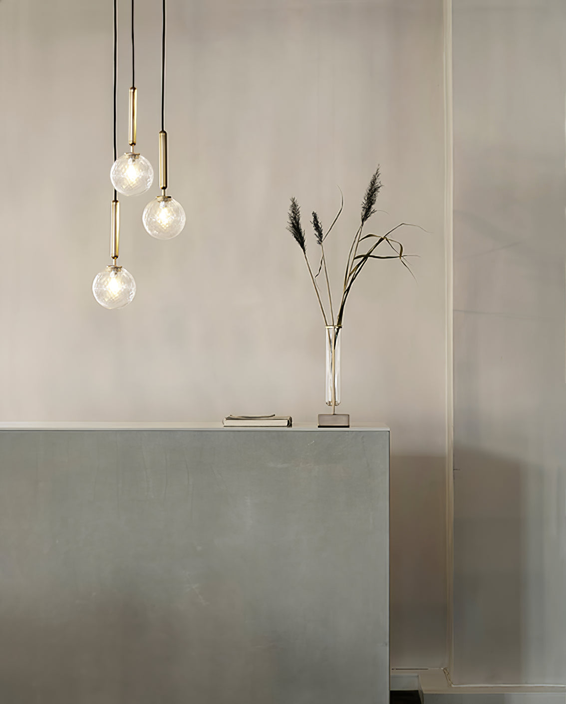 Three Clérachic pendants with clear textured globes on brass rods hang above a stone console and a stem of grass.