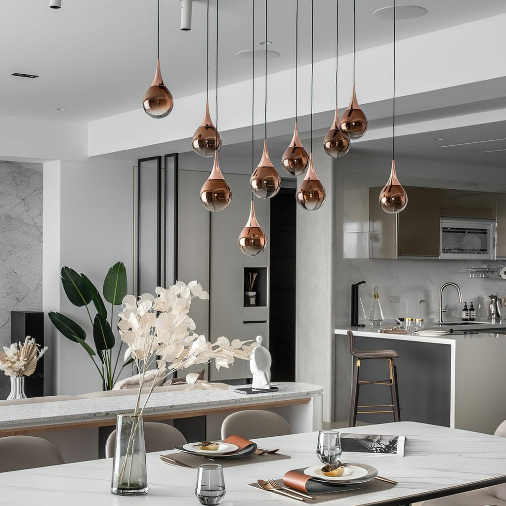 A cluster of rose gold Clérachic pendants hangs at different heights over a white dining table, with a kitchen island behind.