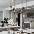 A cluster of rose gold Clérachic pendants hangs at different heights over a white dining table, with a kitchen island behind.