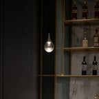 A single Clérachic pendant with a clear teardrop shade glows against a dark wall of shelves holding bottles.
