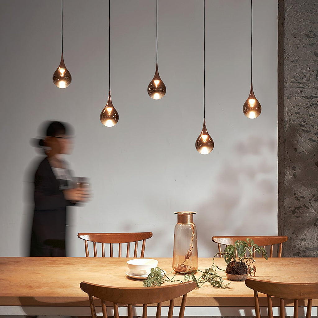 Five Clérachic pendants in rose gold glass hang above a pine dining table, with a blurred figure walking past.