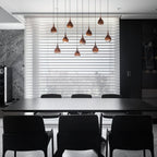 Rose gold Clérachic pendants hang in a staggered group in front of a blinds-covered window, above a black dining table.