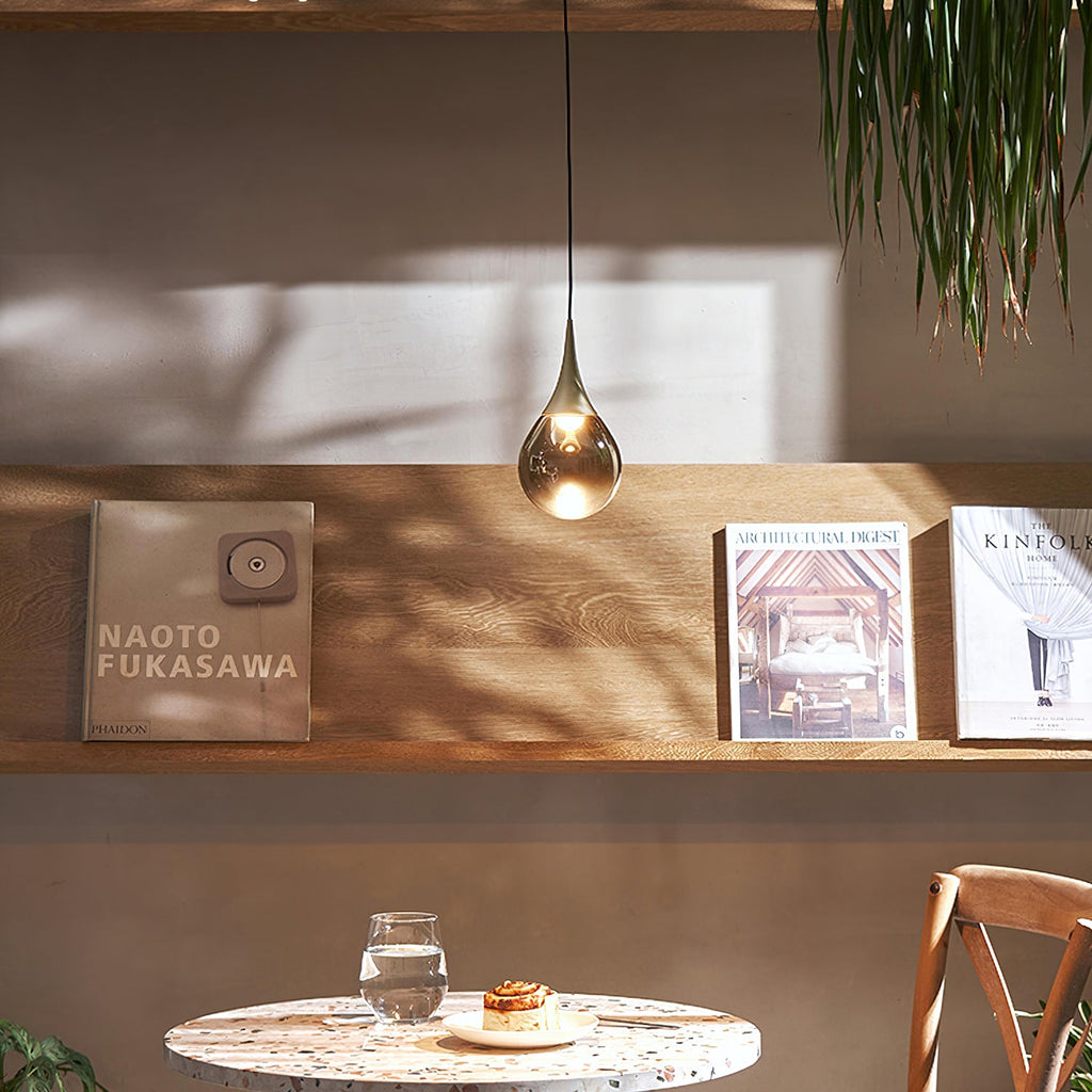 A single Clérachic pendant with a smoky teardrop shade glows above a wooden shelf of books, with a small table below.