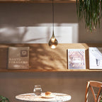 A single Clérachic pendant with a smoky teardrop shade glows above a wooden shelf of books, with a small table below.