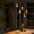 Four warmly lit Clérachic pendants glow in a dim dining room above a wooden table, with tall windows behind.