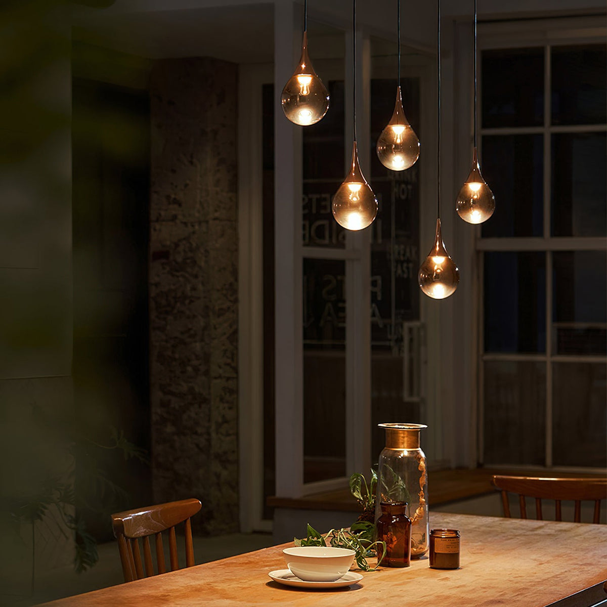 Four warmly lit Clérachic pendants glow in a dim dining room above a wooden table, with tall windows behind.