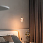 A single Clérachic pendant glows beside a bed with brown curtains and a cream headboard, in a softly lit bedroom.