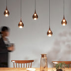 Four Clérachic pendants with rose gold teardrop shades hang in a row above a wooden table and spindle-back chairs.