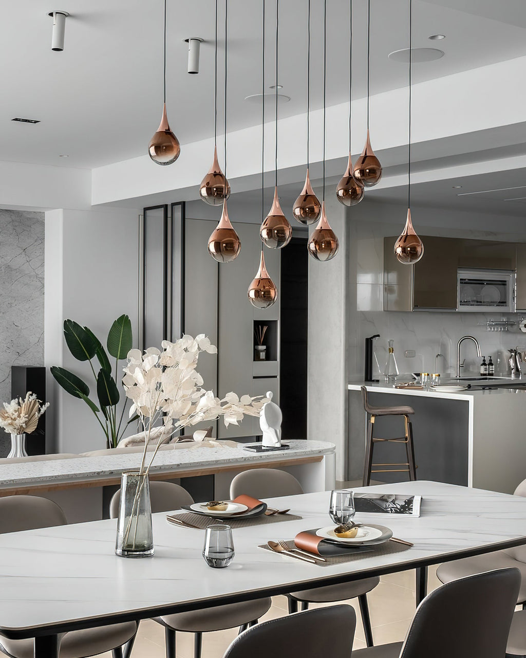 Rose gold Clérachic pendants hang at staggered heights above a white dining table, with a modern kitchen beyond.