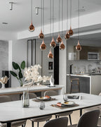 Rose gold Clérachic pendants hang at staggered heights above a white dining table, with a modern kitchen beyond.