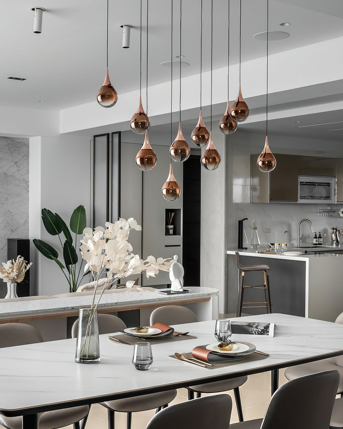 Rose gold Clérachic pendants hang at staggered heights above a white dining table, with a modern kitchen beyond.