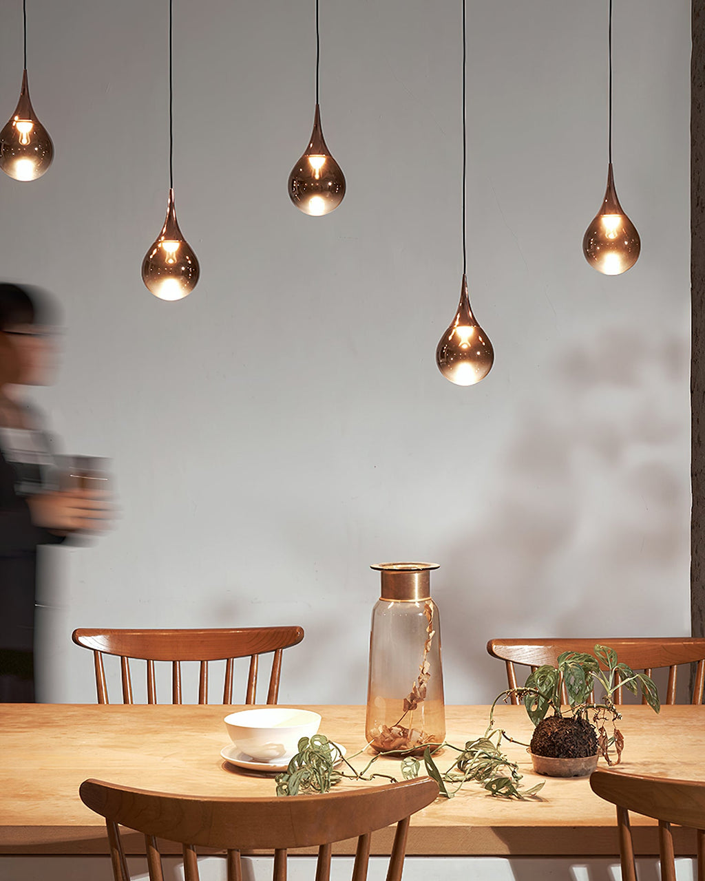 Four rose gold Clérachic pendants hang in a row above a pine table with a glass vase, beside a blurred passer-by.