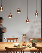 Four rose gold Clérachic pendants hang in a row above a pine table with a glass vase, beside a blurred passer-by.