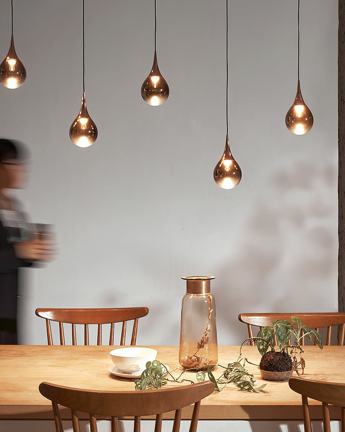 Four rose gold Clérachic pendants hang in a row above a pine table with a glass vase, beside a blurred passer-by.