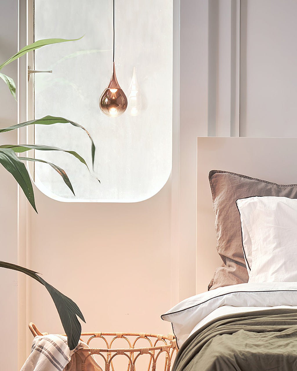 A single rose gold Clérachic pendant hangs beside a rounded window above a bed with grey-green linen and a wicker basket.