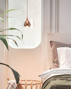A single rose gold Clérachic pendant hangs beside a rounded window above a bed with grey-green linen and a wicker basket.