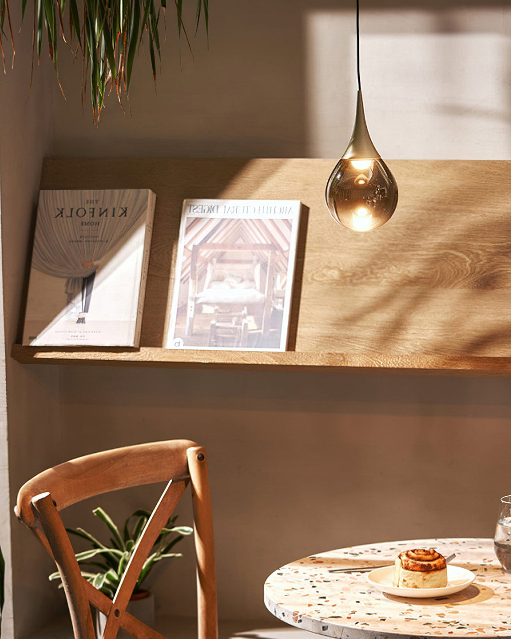 A Clérachic pendant with a smoky teardrop shade hangs by a wooden shelf of magazines, above a speckled round table.