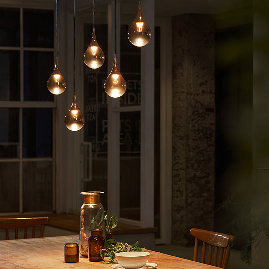 Rose gold Clérachic pendants glow above a dark dining table with candles, a glass vase and a bowl, in a dim room.