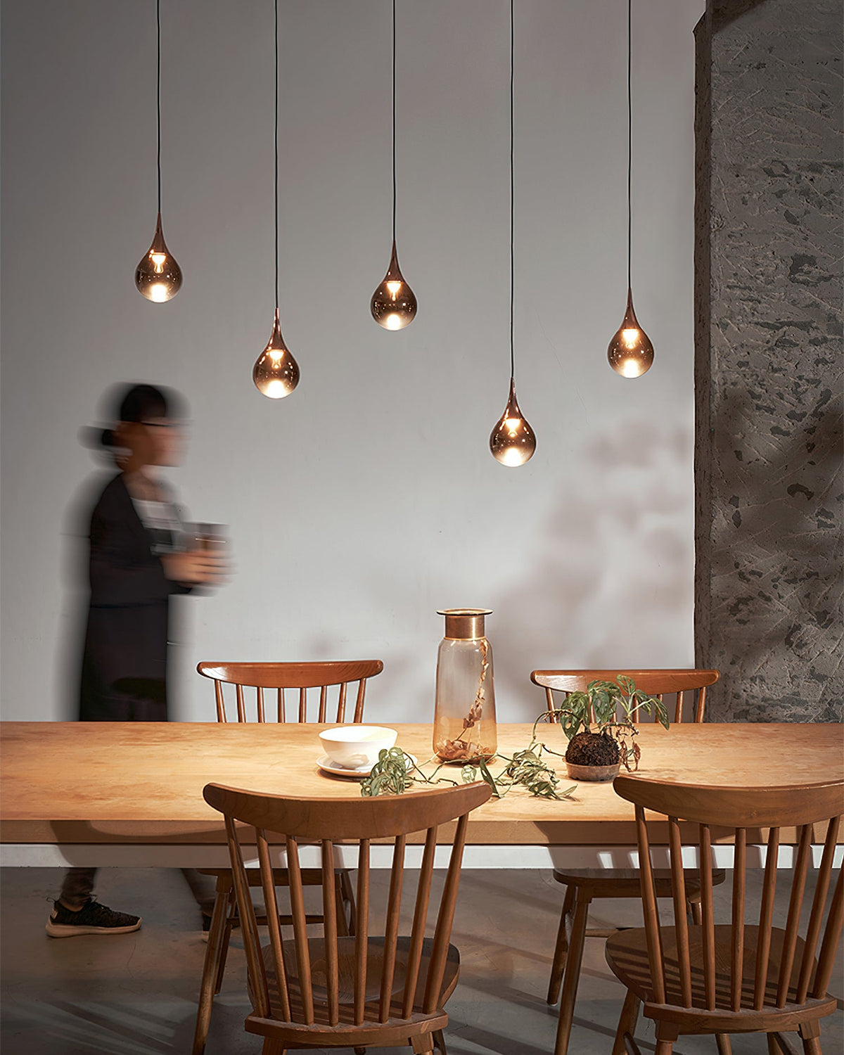 Rose gold Clérachic pendants in a staggered row above a wooden table, with a figure in a dark jacket moving past.