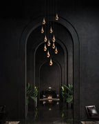 A tall cluster of rose gold Clérachic pendants hangs down a dark arched hallway, with green plants and a seat far beyond.